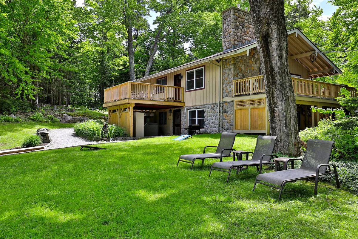 A wide exterior shot of the house with an expansive lawn, a large deck, and surrounding trees. The deck is set up for outdoor dining or relaxation, seamlessly connecting the home to its natural surroundings and offering space for games or morning coffee.
