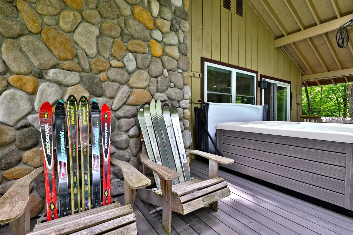 An exterior scene with a bubbling hot tub set against a stone wall, flanked by wooden chairs and winter skis. The space feels inviting and cozy, with the suggestion of sunset views and après-ski relaxation outdoors.