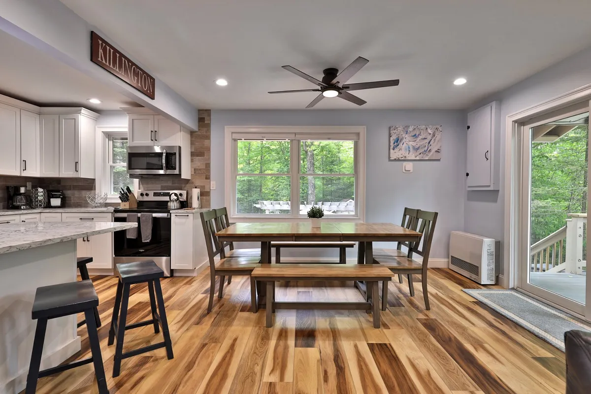 A bright, open dining and kitchen area with a large wooden dining table set by expansive windows, a granite-topped kitchen island with bar seating, and stainless steel appliances. The room exudes warmth and is clearly designed for group meals and gatherings.