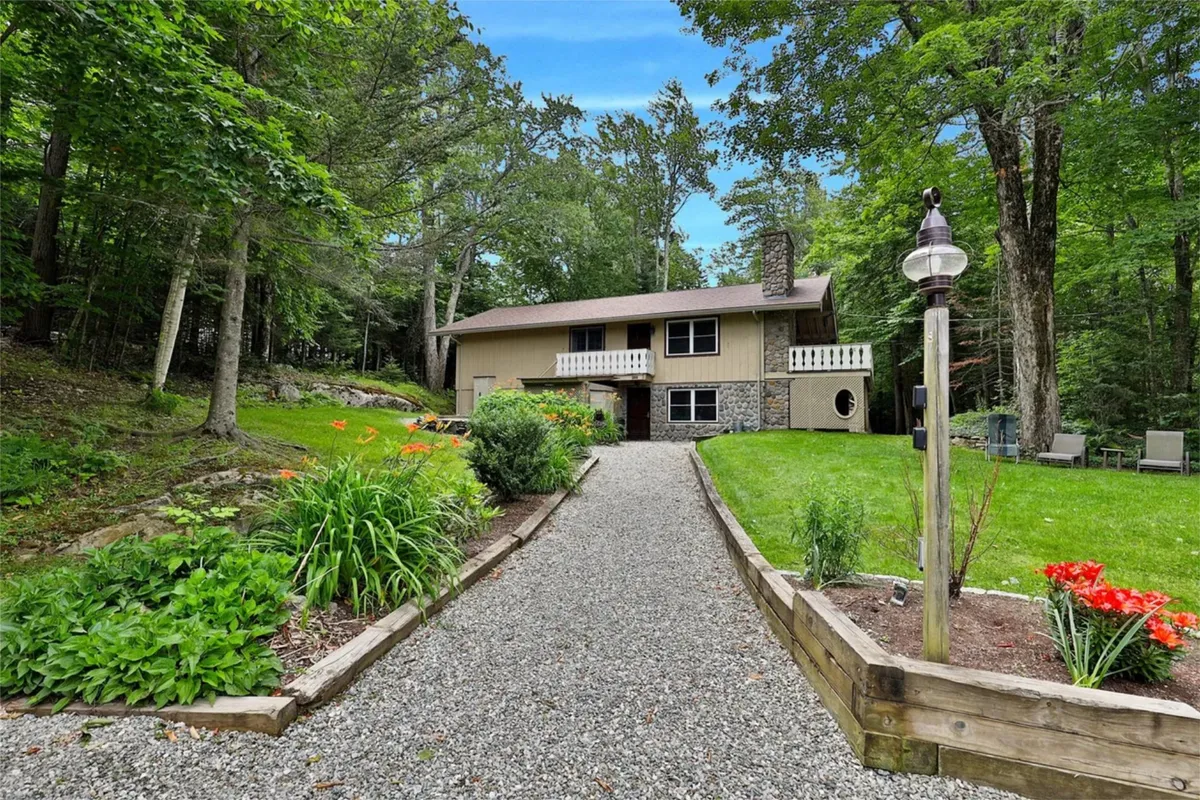 The image showcases the exterior of a newly renovated ski home nestled among tall trees, with a welcoming front porch, a tidy gravel pathway, and a beautifully landscaped garden. The setting is tranquil, surrounded by nature, evoking a sense of peaceful retreat and offering glimpses of where guests might relax, gather outdoors, and enjoy the fresh Vermont air.