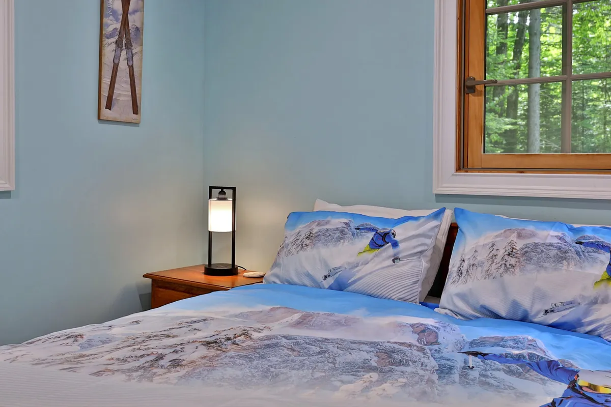 A cozy bedroom featuring a bed with mountain-themed bedding, a bedside lamp, and a window offering views of the surrounding trees. The mood feels calm and inviting, perfect for winding down after a day of adventure.