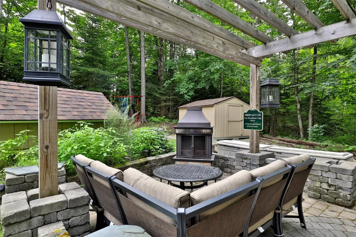 A cozy stone fire pit surrounded by comfortable seating under a wooden pergola, framed by lush greenery. The scene is perfect for family gatherings, relaxing evenings by the fire, and sharing stories under the stars.