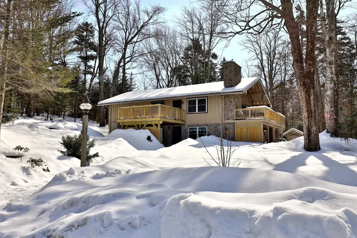 A charming wooden house with a stone chimney sits nestled in a snow-covered landscape, with a balcony overlooking the wintry scene. The setting suggests an inviting retreat after a day of winter sports, with ample space for family gatherings and cozy evenings by the fire.