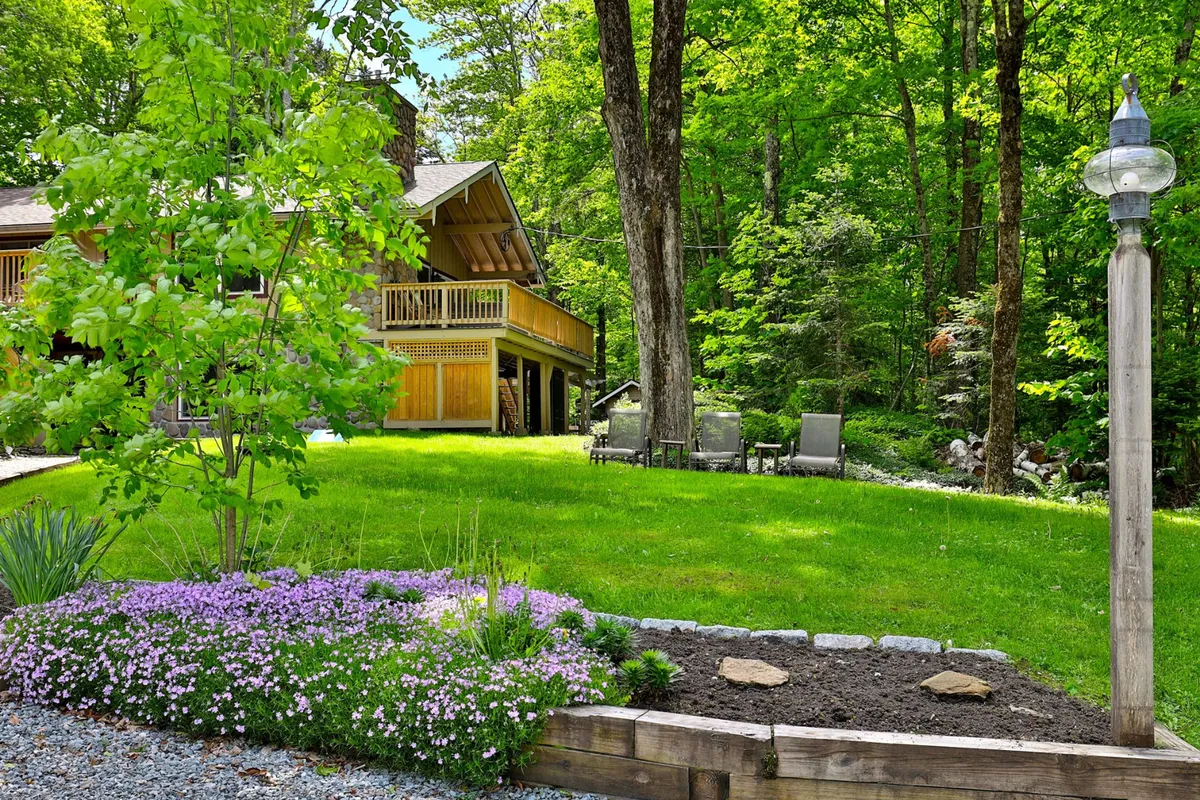 An inviting backyard scene featuring a wooden deck with lounge chairs, green grass, a colorful flower bed, and a lamp post. The deck area looks out onto a tranquil natural setting, suggesting a relaxing retreat surrounded by lush greenery.