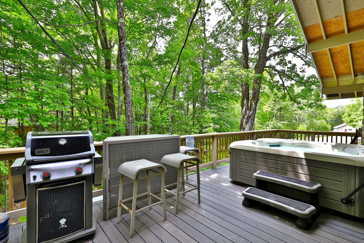 A spacious outdoor area featuring a grill, bar stools, and a bubbling hot tub nestled among surrounding trees. The setup is ideal for socializing, unwinding, or celebrating under the stars, aligning with the notion of resort-like amenities at home.